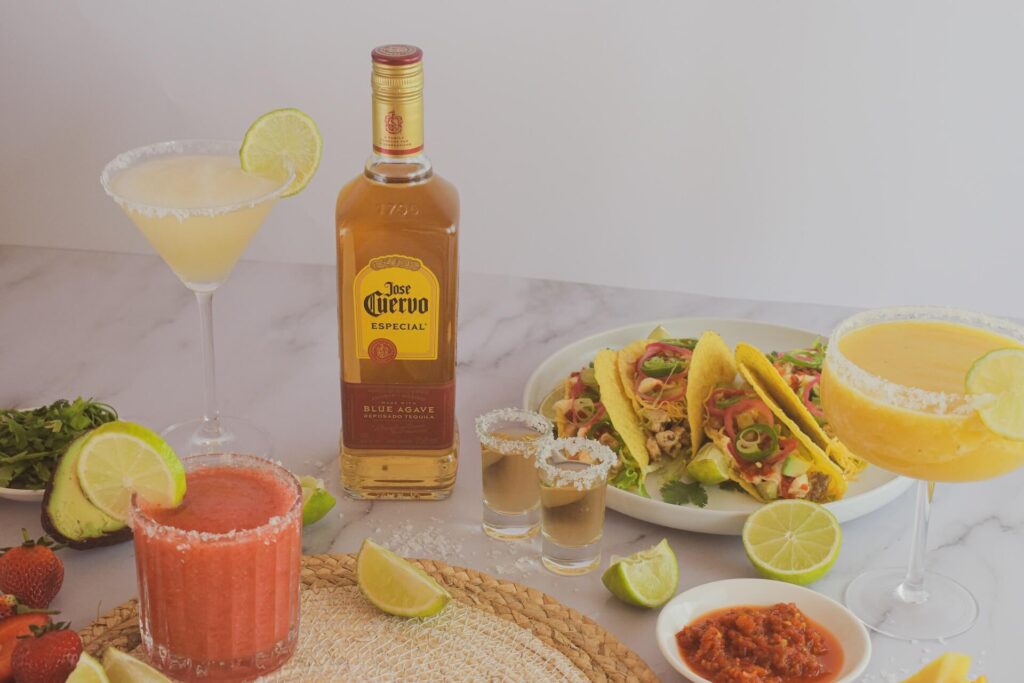 A mexican food dinner is set up on a marble counter with a white wall in the background. A plate contains four overflowing tacos, while beside it is a bowl of red salsa and two full shot glasses with salt rims. A golden bottle of Jose Cuervo sits beside it. Yellow and pink margaritas sit in various styled glasses around the food.