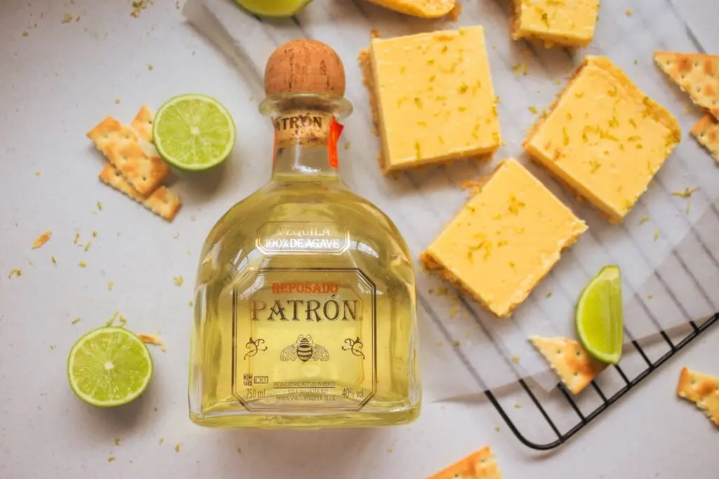 A yellow bottle of Patron sits in the near middle of the image. Behind it is a white counter top with yellow dessert bars, slices of lime and saltine crackers.
