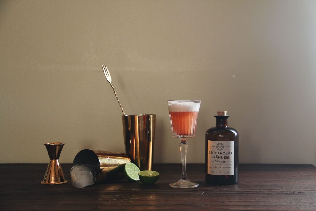 A cocktail making kit sits on a wooden table. It contains a shot glass, two shakes, sliced limes, a pink foamy cocktail and a brown bottle of dry gin.
