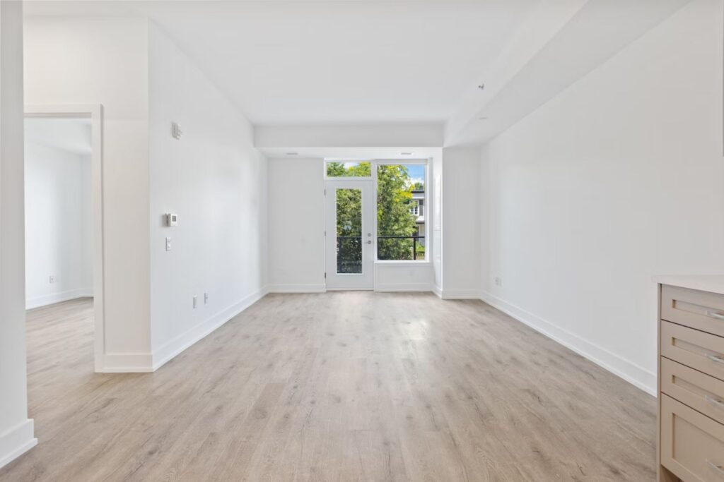 Image is of an empty apartment with light wood floors and white walls, with a door leading to some trees outside. Some first apartment essentials would make it feel more lived-in and cozy.