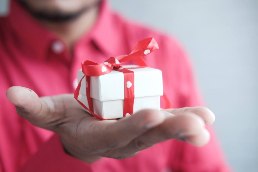 A man in a red button-down shirt holds out a small white box in one of his hands. The box is wrapped in red ribbon with white hearts on it.