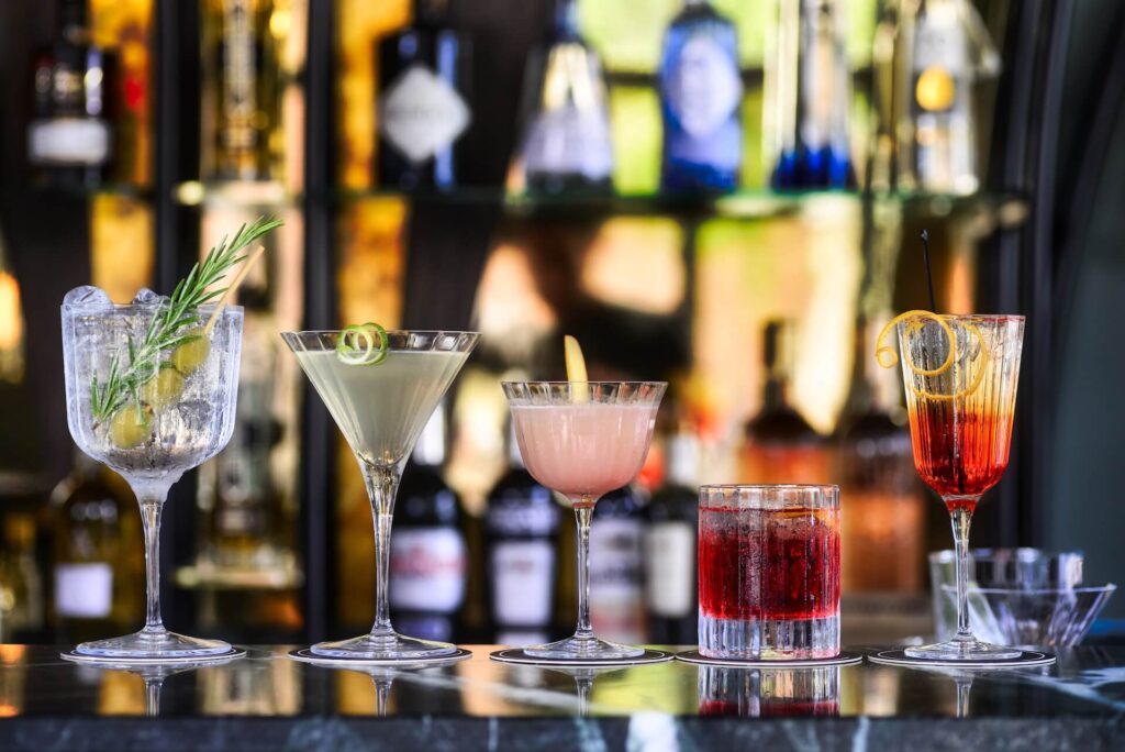 Five cocktail glasses of different shapes and heights sit next to each other on a black marble counter. Bottles of alcohol are on blurred shelves in the background. The cocktails are clear, gray, pink and red. Four have different garnishes.