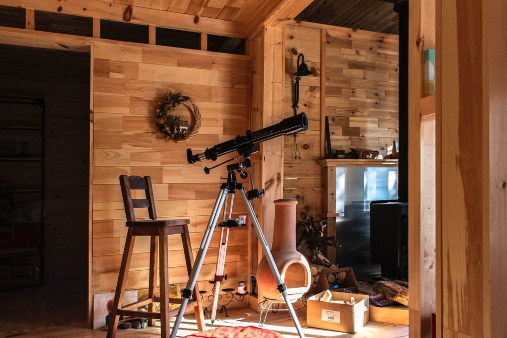 A wooden cabin with two hallways. A large telescope sits on a tripod stand in front of a large wooden chair. Various household decor are scattered on the floor like someone just moved in. A brown wreath with dark foliage hangs on the wall behind the telescope.