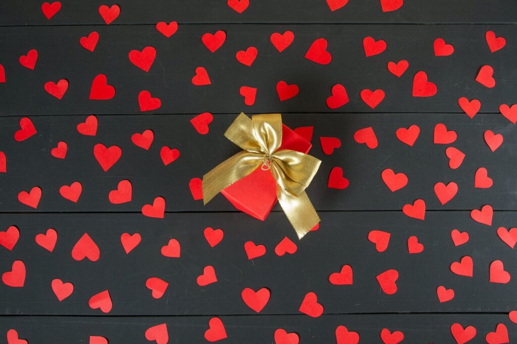 A rectangular red box sits on a floor of dark brown boards. A gold bow is tied around the box and red confetti hearts cover the floor boards.