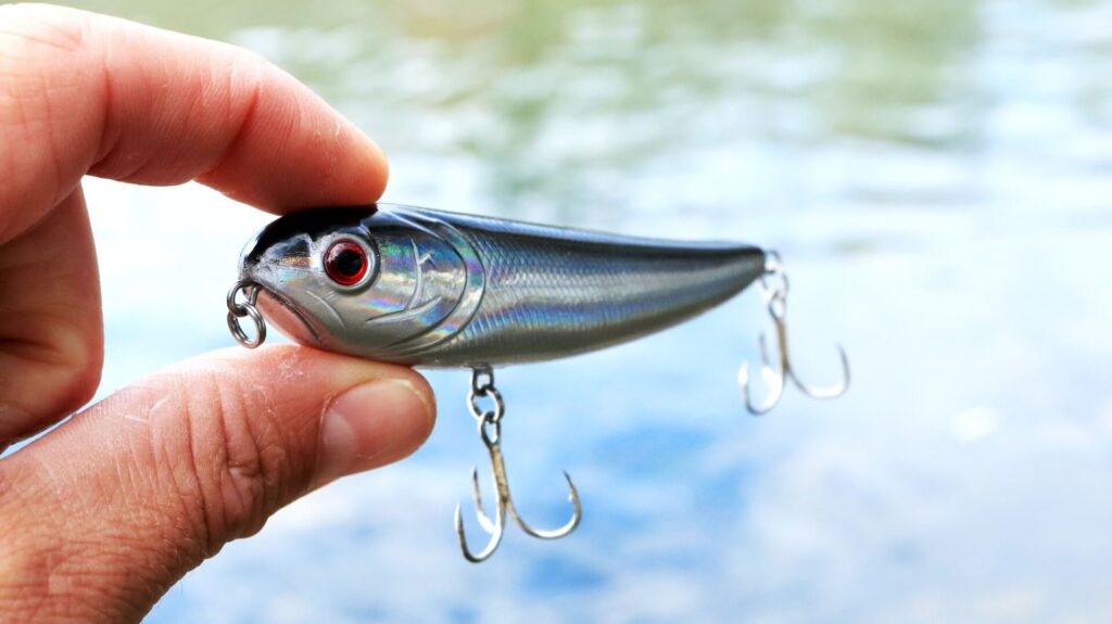 A person holds a small fishing lure up to the camera. Only three fingers and the lure are in focus. The lure is shaped like a small silver fish with red eyes. Two three-pronged hooks hang off both ends. The blurry background is rippling water.