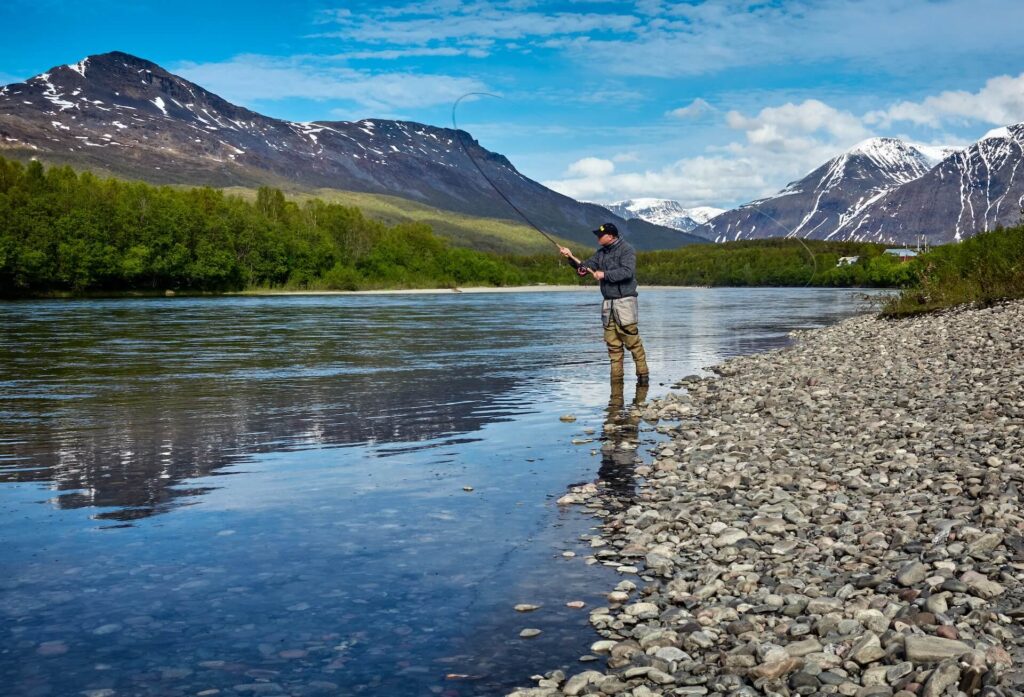A man stands with his boots in a clear river. He wears long green pants, a long-sleeved blue shirt and a black baseball cap. He's mid-cast with a long black fishing pole. The shore is made of small rocks. A gray mountain topped with white snow is across the background. Green trees are below it and blue sky with big white clouds is above it.