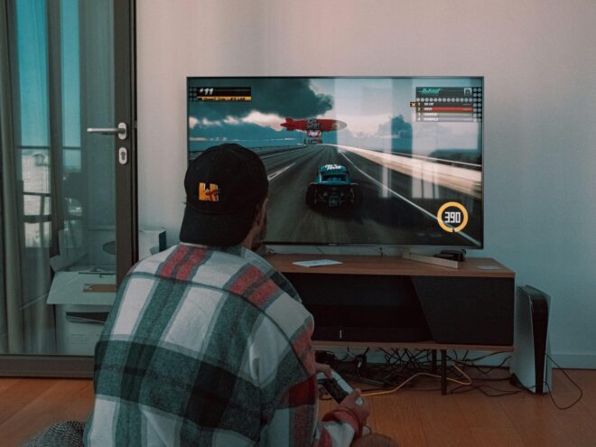 A person playing a racing video game on a large flat-screen TV in a living room. The screen shows a car driving on a racetrack with a speedometer indicating 390 km/h.
