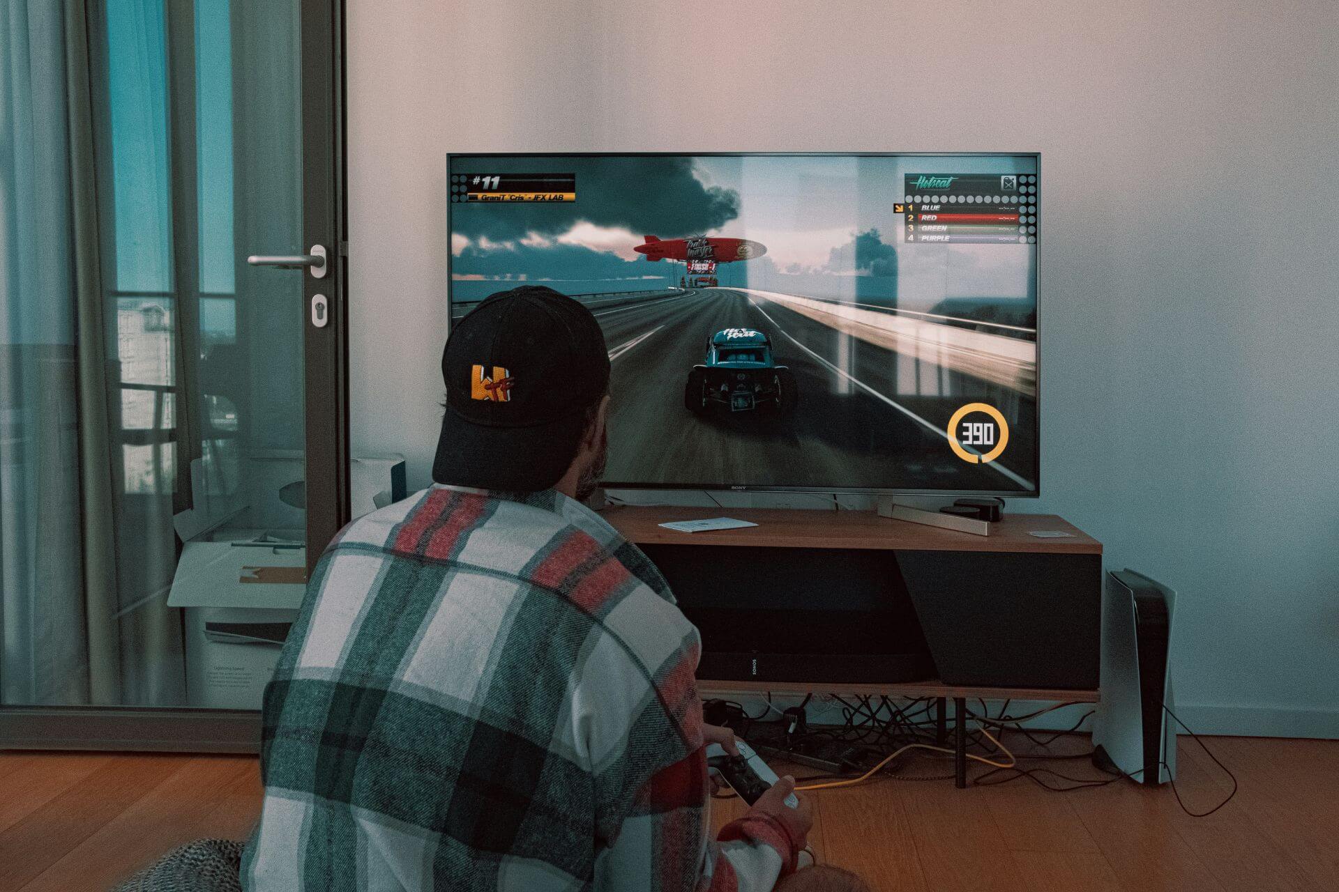 A person playing a racing video game on a large flat-screen TV in a living room. The screen shows a car driving on a racetrack with a speedometer indicating 390 km/h.