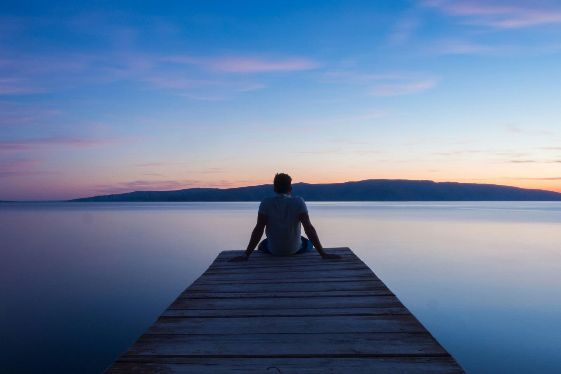 A person sitting at the end of a wooden dock looking out at a calm lake with hills in the background and a colorful sunset sky.