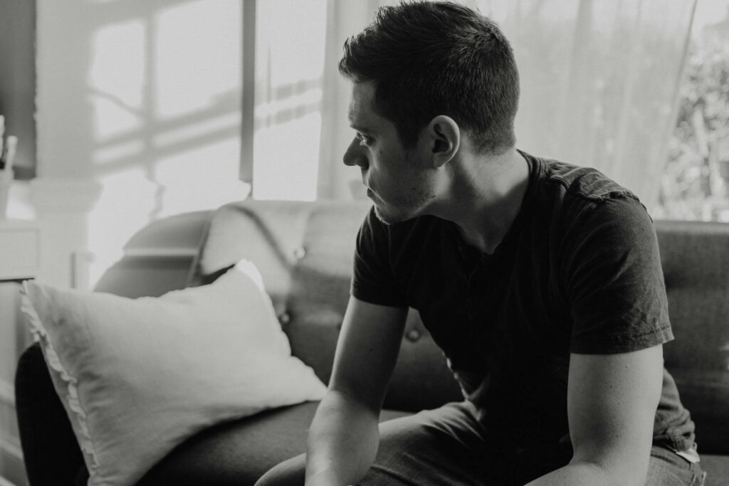 A person in a dark shirt sitting on a couch with their body turned slightly to the side, resting an elbow on the back of the couch. There is a pillow next to them and a bright window in the background. The image is monochrome.