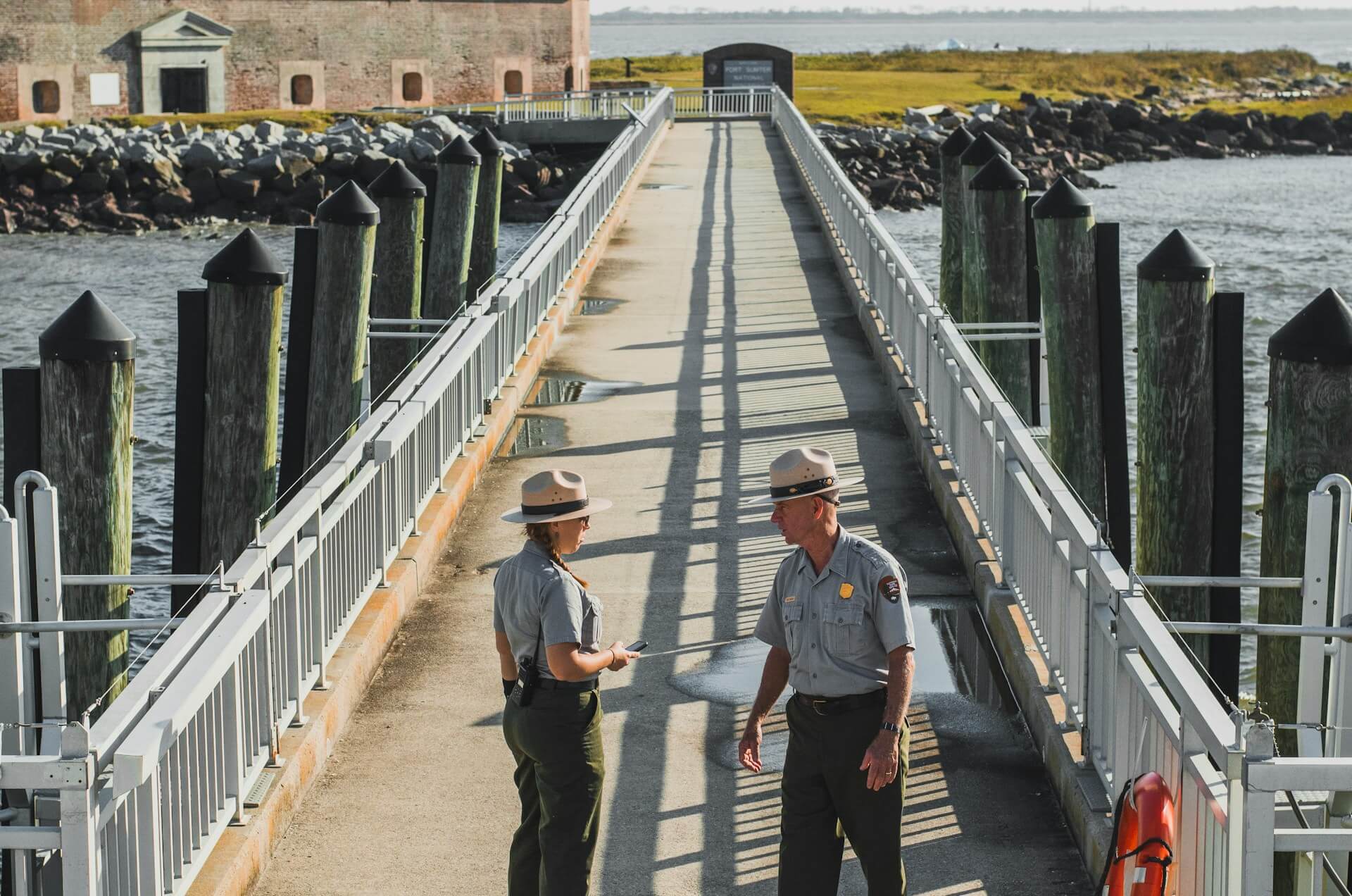 Two park rangers talk on a bridge spanning shallow water and leading to a historical building in the distance. It's likely a fort. A large inlet is behind the building in the distance.